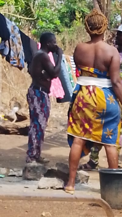 A man with no shirt and patterned pants stands on a rock, hand near his face, next to a clothesline with assorted garments. To the right, a woman with intricate braided hair wears a blue and yellow strapless dress with star and flower designs. A man in camouflage pants stands partly behind her, and a black bucket is in the foreground. The scene appears to be outdoors in a somewhat rural or impoverished setting.