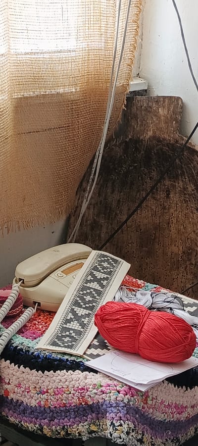 The image shows a still life scene. A landline telephone sits on a colorful, textured fabric. Adjacent to the telephone is a paper pattern with a geometric design. A ball of red yarn rests nearby. The background includes a burlap curtain and a dark, wooden object. The specific location within Кемуль, Russia, cannot be determined from this image alone.