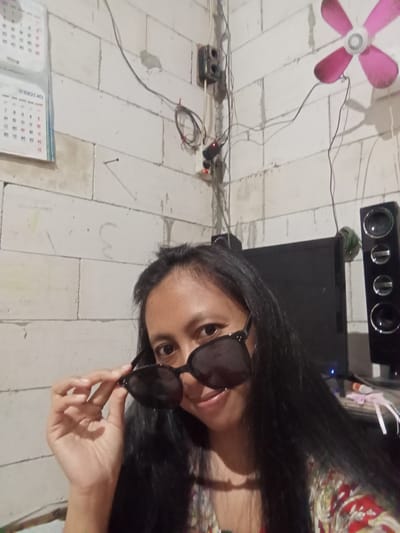 The image is a selfie taken by a woman holding sunglasses in front of her face. She has long, dark hair and is wearing a patterned top. Behind her, there is a television, speakers, a pink electric fan and a power strip. The wall is made of white rectangular blocks. A calendar hangs on the wall. The location is Baron, Indonesia.