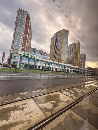 The image shows a street view of modern residential buildings and a shopping mall, with tram tracks in the foreground. The buildings are tall and multi-story, with a mix of glass and brick facades. The shopping mall, named "ПАРК МОЛЛ" (PARK MALL), has a green and white exterior with large windows and visible store signage. Some of the store names visible include "ИНГОСПОЛИС" (INGOPOLIS) and "Супермаркет" (Supermarket).

The scene appears to be set in Sinyavino, Russia. The time of day is unclear, but the sky is overcast with some areas of bright light, suggesting it could be late afternoon or a cloudy day. The ground near the tram tracks is wet, indicating recent rain or damp conditions. There are a few people visible walking near the entrance of the shopping mall. The overall mood is neutral, depicting an urban environment. The image is oriented with the top of the buildings appearing at the bottom of the frame, suggesting it may have been taken from a moving vehicle or a similar perspective.