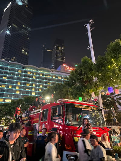 The image depicts a nighttime scene in Central Jakarta, Indonesia, likely near the Hotel Indonesia Kempinski, as indicated by the prominent hotel sign. A bright red fire truck is the central focus, parked on a street or plaza. A crowd of people, including men, women, and children, are gathered around and on the fire truck, many appearing to be spectators or participants in an event. Some individuals are seated on the truck's exterior, while others stand nearby. The fire truck itself has text on its side, including "MEDIUM PRESSURE PUMP" and "GAMBIR CREW". In the background, towering skyscrapers and illuminated buildings are visible, characteristic of a city at night. Trees adorned with string lights and colorful lanterns are also present, adding to the festive atmosphere. The lighting suggests it is after dark, with artificial lights from buildings, the fire truck, and decorative lighting illuminating the scene. The overall impression is of a public gathering or celebration taking place in an urban setting.