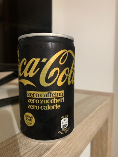 A close-up, horizontally oriented, slightly low-angle shot shows a can of Coca-Cola Zero in black and gold packaging. The can is positioned on a light-colored, textured surface, possibly a wooden shelf or countertop. The background is a plain, off-white wall.

The can features the iconic Coca-Cola logo in a prominent gold script. Text on the can indicates "zero caffeina", "zero zuccheri", and "zero calorie", all in gold lettering. There's also a yellow oval sticker with the text "nuovo look" and "STESSO GUSTO" (new look, same taste). Nutritional information is visible on the right side of the can, stating "150 ml", "1.4 kJ / 0.3 kcal", and "0%".

The lighting appears to be artificial and casts subtle shadows. The can shows some condensation, suggesting it is chilled. The overall scene is simple and focused on the product. The image does not contain people or depict a specific event, but rather showcases the product in a static setting.
