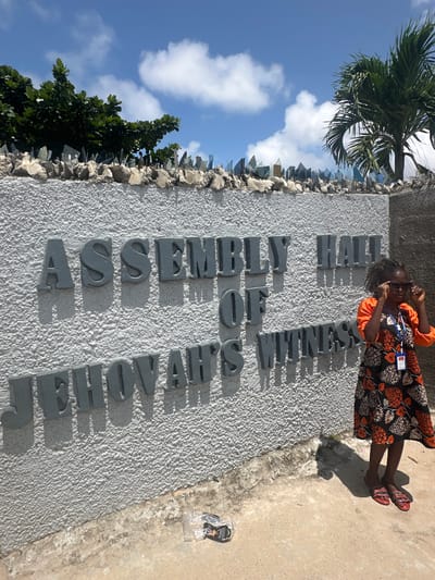 The image captures an outdoor scene at the Assembly Hall of Jehovah's Witnesses in Itoga, Nigeria. A young girl, wearing sunglasses, an orange shirt, and a patterned dress, stands to the right of a textured white wall. The wall features prominent grey lettering spelling out "ASSEMBLY HALL OF JEHOVAH'S WITNESSES." The top of the wall is lined with what appear to be shards of glass or broken material, set into a concrete barrier, likely for security. Behind the wall, lush green trees and a palm tree are visible against a bright blue sky with scattered white clouds. The ground in front of the wall is a rough, light-colored concrete or dirt surface, with some debris, including a crumpled plastic bag, on the lower right. The sunlight casts distinct shadows from the lettering and the girl onto the wall and ground, indicating it is a sunny day.