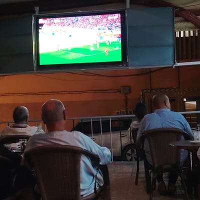 In Ouaouizert, Morocco, four individuals are seated in what appears to be an outdoor seating area, facing a large screen displaying a soccer match. The screen is mounted above and to the left of the seated patrons. The environment suggests a casual setting, possibly a cafe or restaurant, with patrons seated in wicker chairs.

In the foreground, three men are visible from behind. The man on the left is wearing a light-colored shirt with vertical stripes. The man in the center is bald and wearing a light-colored shirt. The man on the right is also bald and wearing a light blue collared shirt. A fourth person, whose back is turned to the camera, is partially visible in the far right of the frame, wearing a white shirt.

The screen shows a bright green soccer field with players in white and orange uniforms and a densely packed audience in the background. Behind the seated patrons, a silver car is visible, separated by a metal barrier. The walls of the building are painted an orange hue, and electrical conduits are visible running along the wall. The floor appears to be paved. The lighting suggests it is daytime, but the area might be shaded.