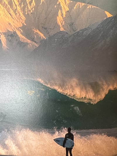 The image depicts a surfer in silhouette, holding a surfboard, standing in the shallow, foamy water at the edge of a large wave. The wave is illuminated by golden light, suggesting either sunrise or sunset, and it is breaking with spray. In the background, a mountain range with sharp, angular peaks is visible, bathed in the same warm light. The sky above the mountains is a hazy grey. The location is identified as Ahangama, Sri Lanka, known for its surf breaks. The scene is a dynamic capture of a surfer preparing to ride a powerful wave against a dramatic mountainous backdrop.