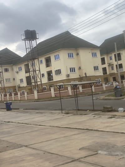 The image shows a multi-story building in Iddo Tudunwada, Nigeria. The building has a light-yellow exterior and numerous windows with blue frames. A dark green roof is partially visible. Power lines run across the top left of the frame. A water tank supported by a metal structure is seen adjacent to the building. A street runs alongside the building, and a concrete walkway is in the foreground. Several bins are visible. The sky is overcast.