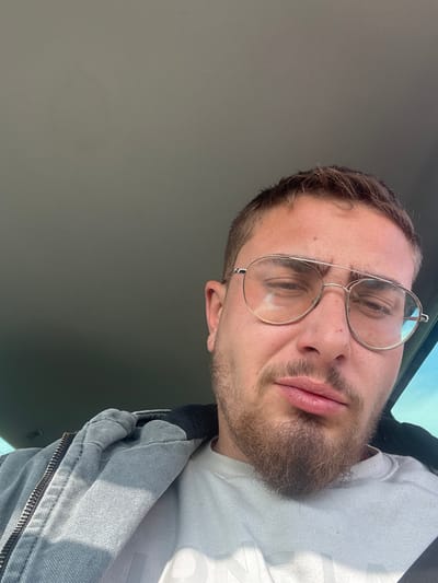 The image presents a close-up, upward-angled portrait of an adult male, likely a selfie, captured inside what appears to be a vehicle. The subject has fair skin, short light brown hair, and a light brown beard and mustache. He wears large, wire-rimmed aviator-style glasses, with reflections visible on the lenses. His eyes are partially closed or squinting, and his lips are slightly pursed. He is dressed in a light grey zippered hoodie over a lighter-colored long-sleeved shirt, with a portion of a graphic or text visible on the shirt's chest. The interior roof lining of the vehicle, light grey in color, occupies the upper background. A faint blue hue, indicative of an exterior sky or window, is visible on the far right side of the frame. No discernible landmarks, signs, or contextual elements within the image provide information to identify the specific location as Manastir, Bulgaria, or any particular city area.