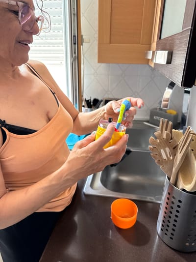 A woman wearing glasses and an orange top is in a kitchen. She is holding a yellow object with a blue and green attachment. The kitchen has wooden cabinets, a stainless steel sink with wooden utensils in a container next to it, and an orange cup. The location is Aprilia, Italy.