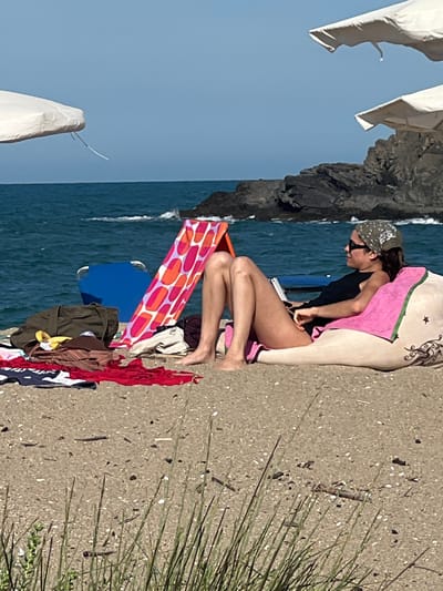 A woman is relaxing on a beanbag on a sandy beach, facing to the right. She is wearing a black tank top and a bandana. The beanbag is pink and white with some decorative elements. In front of her, there are various beach items, including towels, a bag, and a foldable blue chair. A brightly colored towel with an orange and pink pattern stands upright behind some of the items. The sea is visible in the background with some rocks. The sky is clear and blue. The location is the beach in Sinemorets, Bulgaria.