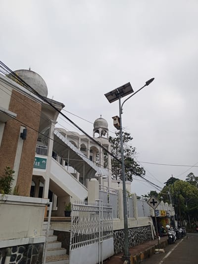 A low-angle, full shot captures a street scene under an overcast sky. On the left, a section of a building with a beige textured facade and a white domed roof is visible, with black power lines strung across it. Adjacent to this building, a white staircase with a metal canopy leads upwards, with another white domed structure visible in the background. A solar-powered street light with two panels stands prominently in the foreground, equipped with a small, rusty box attached to its pole.

The street itself is lined with a low white fence and a stone wall. Further down the street, a series of shops are visible, with several motorbikes parked along the sidewalk. A pedestrian crossing sign with a black cross on a white diamond is posted near the shops. Trees and lush greenery border the street in the background. The road is paved, and the scene appears to be in a residential or commercial area. No people are clearly depicted in the image. The overall impression is one of a typical urban streetscape on a cloudy day, with religious architecture hinting at a mosque.