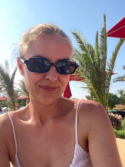 The image shows a woman wearing sunglasses and a white top, possibly a swimsuit. She is outdoors, likely on vacation, with palm trees visible in the background. There are red awnings or umbrellas also visible. Considering the prompt specifies Monastir, Tunisia, it is reasonable to conclude that the woman is at a resort or similar location in Monastir.