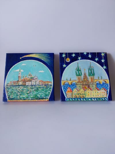 Two small, square paintings are displayed side-by-side on a light gray surface. The painting on the left depicts a cityscape that resembles Venice, Italy, with buildings featuring domed roofs and tall spires situated along a body of water. A yellow and orange shooting star arcs across a dark blue sky above the scene. The painting on the right portrays a scene reminiscent of Prague, Czech Republic, with a prominent bridge connecting two sides of a river. The foreground features colorful, row houses. This painting also has a dark blue sky background, but it is adorned with hanging stars and a yellow circular object, possibly the moon or a lantern. The paintings are not located in the city of Rivne, Ukraine.