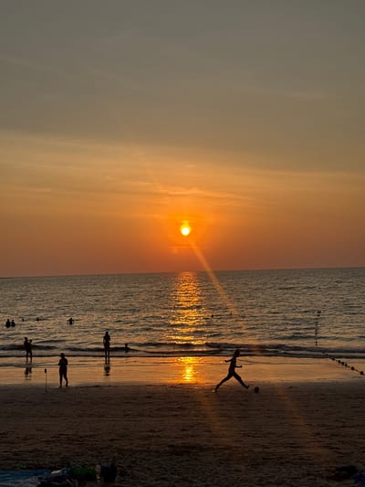 This image depicts a sunset scene at Kamala Beach in Kamala, Thailand, facing west towards the Andaman Sea. The sky transitions from a light grayish-blue at the top to an intense orange-yellow at the horizon. A prominent, bright orange sun is positioned slightly left of center on the horizon, casting a strong, shimmering golden reflection path across the water surface towards the viewer. The sea exhibits gentle waves.
Approximately ten individuals are visible, primarily in silhouette. In the mid-ground, a line of dark buoys extends into the water from the right. Several people are in the water, some swimming and others standing partially submerged. On the wet sandy beach in the mid-ground, one person is captured in motion, mid-stride, kicking a football. Two additional silhouetted figures stand near the waterline; one appears to be holding a dark, elongated object. In the lower foreground, on the darker, drier sand, a blue mat or towel, several bags, and a plastic water bottle are visible.