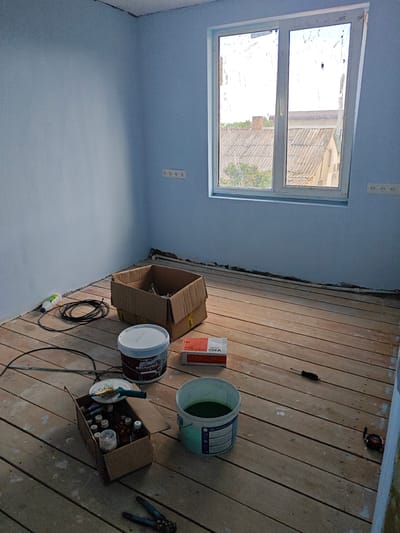 The image depicts an interior room undergoing renovation, likely a bedroom or living area, in a residential building located in the Береговой сельский совет, Ukraine. The walls are painted a light blue color, and the floor is made of unfinished wooden planks. A large white-framed window with two panes is visible, offering a view of rooftops and trees outside. The window has protective film or tape that is partially peeled off.
Several construction and painting supplies are scattered across the floor. In the center-left of the frame, a cardboard box contains various small bottles, possibly paint or sealant. Next to it, a white bucket with a red and white label and a blue lid sits on the floor, along with another bucket containing a dark green liquid. A red and white box, labeled "V-KO", is also present.
To the left of the box, a black electrical cord is coiled, and a white bottle with a green cap is lying on its side. Further left, a tape measure is laid out on the floor. In the foreground, a pair of wire cutters are visible. On the floor, near the window, a screwdriver is placed. Two electrical outlets are mounted on the wall to the right of the window. The overall scene suggests a paused construction or renovation project, with materials and tools laid out in preparation for or during work. No individuals are present in the image.