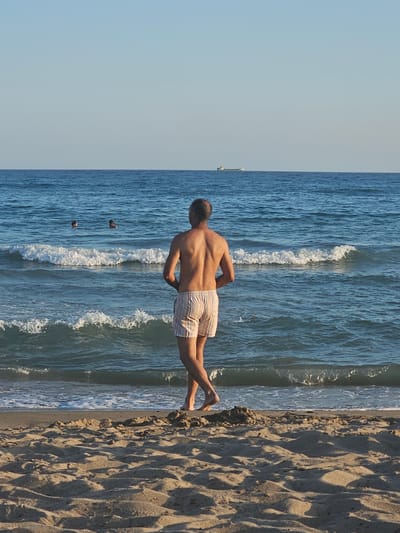 A man in striped swim trunks stands on a sandy beach facing the ocean. The ocean is blue with moderate waves and two people are visible swimming in the distance. The sky is clear and blue. A cargo ship is visible on the horizon. The scene appears to be in the city of Castelldefels, Spain, known for its long sandy beaches and views of the Mediterranean Sea.