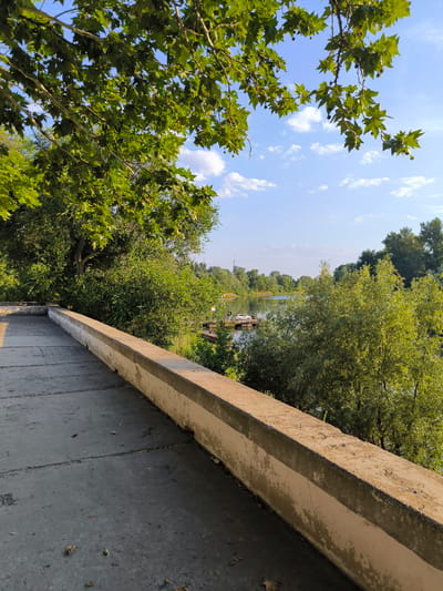 A scenic view of a river on a sunny day. A concrete path with a low wall runs along the left side of the frame, partially obscuring the view of a lush green riverbank. Trees and vegetation cover the landscape, with a few scattered clouds in the bright blue sky. On the river, a small dock with a white car is visible. The overall mood is peaceful and serene, suggesting a quiet moment in nature. The time of day appears to be morning or late afternoon due to the soft sunlight casting shadows on the path. The location is Tiraspol, Moldova.