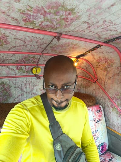 A bald African man with a goatee, wearing glasses and a yellow long-sleeved shirt, poses for a selfie. He is inside a vehicle, possibly a tuk-tuk, with a patterned floral roof and pink metal frame. A brown cross-body bag is slung over his shoulder. A small yellow decorative object hangs from the roof. The vehicle interior appears padded with patterned fabric. A seat is visible to his right.