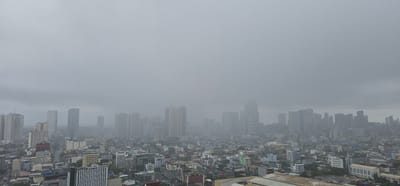 This is a wide, aerial shot of a dense urban landscape under a heavily overcast sky, suggesting a cloudy or foggy day. The city is filled with numerous buildings of varying heights, from mid-rise apartment complexes to tall skyscrapers that dominate the skyline. The architectural style appears to be modern, with many buildings featuring clean lines and glass facades.

In the foreground and mid-ground, a vast expanse of lower-rise buildings forms a dense urban fabric. These structures are a mix of residential and commercial properties, with visible rooftops, windows, and balconies. Some streets are visible, showing cars and traffic, indicating the city is active.

The background is characterized by taller, more modern skyscrapers, some of which are partially obscured by the atmospheric conditions. The overall impression is one of a bustling metropolitan area. The muted color palette, dominated by grays and blues, reinforces the subdued weather and creates a somewhat somber or atmospheric mood.

The scene depicts the everyday activity of a large city, with no specific event or prominent human activity being the focus. The time of day is difficult to ascertain precisely due to the lack of direct sunlight and shadows, but it appears to be daytime. The location is indicated to be Makati, Philippines, a major business district. No visible text is discernible in the image.