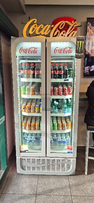 A brightly lit, white, double-door commercial beverage refrigerator stands centrally, fully stocked with various cold drinks. Above the unit, a prominent golden "Coca-Cola" sign with individual letters is mounted, accompanied by a red circular Coca-Cola logo. To the immediate right, a tall, black Monster Energy drink can is depicted on a vertical sign.
The left refrigerator door displays four shelves. From top to bottom, these shelves are stocked with: bottles of Coca-Cola, various Fanta cans (orange, green, yellow, light blue), additional Fanta cans (predominantly orange), and rows of small plastic water bottles with blue labels.
The right refrigerator door also contains four shelves. From top to bottom, these shelves hold: bottles of Coca-Cola and other dark-colored sodas, red Coca-Cola cans, clear or light-colored bottled beverages (possibly Ramune), and assorted Fanta cans (green and orange). Each refrigerator door features a "Coca-Cola" logo near the top. The lower section of the refrigerator is equipped with a grated vent.
The floor is surfaced with light grey square tiles. To the left, a vertical green element, likely a door frame or partition, is visible against a light pink wall. To the right, a person wearing a dark jacket is seated on a dark stool with a silver frame, positioned at a light blue-green counter. Behind the counter, a light beige wall is visible, and a television screen displays a blurred image. The specific location within the city of Villeneuve-d'Ascq, France, cannot be determined from the visual evidence provided.