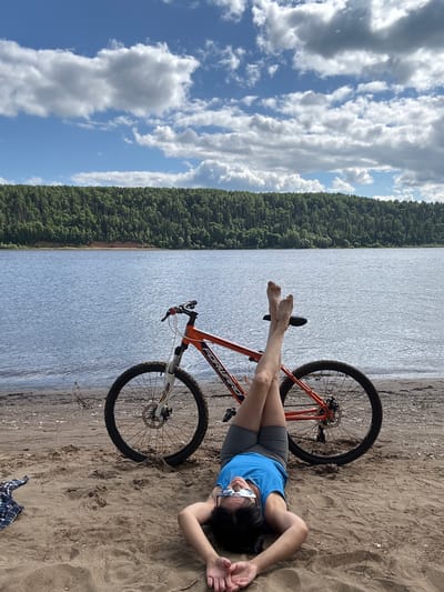 A person lies on their back on a sandy riverbank, with their legs propped up on a bicycle seat. The bicycle is an orange and white mountain bike, positioned upright behind the person. The person is wearing a blue t-shirt, grey shorts, and glasses. They are facing upwards, with their hands resting on their chest. Behind the riverbank is a wide, calm body of water, and across the water is a dense, green forest that covers a hillside. The sky above is blue with scattered white clouds. The scene suggests a leisurely moment by the river. The location is identified as Ol'khovskoye, Russia.