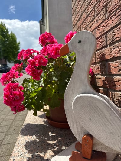 An outdoor, eye-level shot shows a wooden goose decoration with a bright orange beak standing on a granite ledge, next to a pot of vibrant pink geraniums. The goose is positioned to the right of the frame, with its body angled towards the viewer. Behind the flowers and goose, a textured red brick wall creates a backdrop. To the left of the flowers, a paved walkway is visible, and beyond that, a street with parked cars and green trees under a blue sky with scattered clouds. The sunlight casts shadows of the geranium leaves and flowers onto the granite ledge. This scene is located in Bietigheim-Bissingen, Germany.