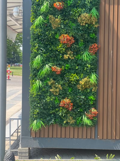 A vertical, artificial plant wall covers the majority of the frame, featuring various shades of green foliage interspersed with clusters of bright orange-red flowers and yellowish-brown sprigs. The plant wall is mounted against a dark grey panel, which is itself set against a background of horizontal brown wooden slats. The overall scene appears to be an outdoor setting, possibly a building exterior or a decorative garden feature. Below the plant wall, a polished, reflective surface mirrors the scene above, showing trees and the sky, as well as a paved walkway with red and white bollards. The lighting suggests it is daytime, likely a sunny day given the bright reflections and shadows. No people are visible in the image. The scene is calm and decorative.