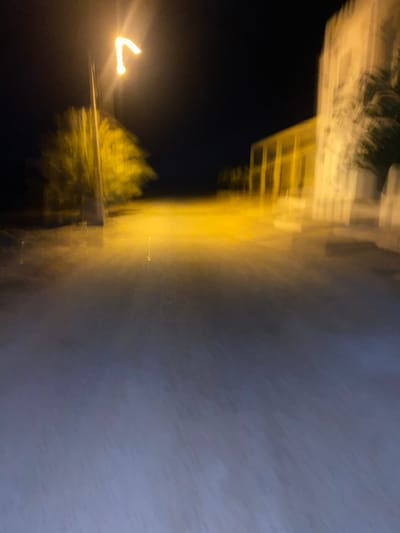 This image depicts a nighttime outdoor scene in the city of الغنادة, Tunisia, characterized by significant motion blur. The primary light source is a bright, yellow-orange streetlamp positioned on the left side of the frame, illuminating a blurred tree immediately to its right. The foreground consists of a dark, flat surface, likely a road or ground, which reflects some of the yellow-orange light, transitioning to a darker, indistinct blue-grey further into the foreground. On the right side of the frame, a multi-story building structure is visible, illuminated by a combination of ambient light and the streetlamp, appearing in blurred shades of yellow-orange and off-white. This structure features vertical elements consistent with columns or arches. No individuals or vehicles are discernible within the scene. The background is uniformly dark, indicating a night sky or unlit distance.