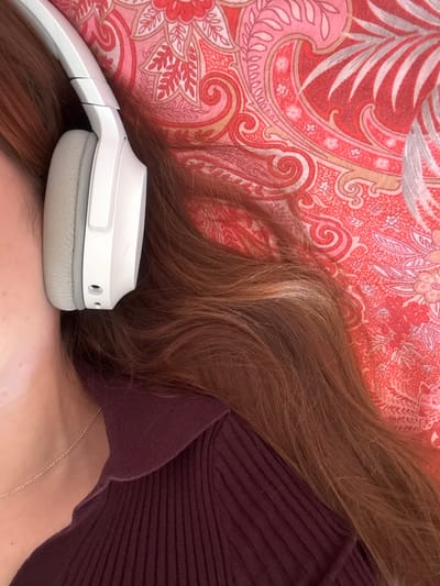 The image displays a close-up, partial view of a person, likely female, lying down, observed from an overhead perspective. The exact location within the city of Visp, Switzerland, cannot be determined from the visual evidence provided. The subject's head, specifically the right side including the ear, jawline, and a significant portion of long, reddish-brown hair with lighter highlights, is visible on the left side of the frame. A white over-ear headphone cup with a light grey ear pad covers the right ear. The headband extends upward and is partially visible. Below the head, the person's upper torso and shoulder are visible, clad in a dark red or maroon ribbed polo-style shirt. A thin, gold-colored chain necklace rests on the skin near the collarbone. The background consists of a fabric with a repeating paisley or floral pattern in shades of red, pink, orange, and beige, likely a pillowcase or blanket. No dynamic actions or interactions beyond the individual wearing headphones are depicted.