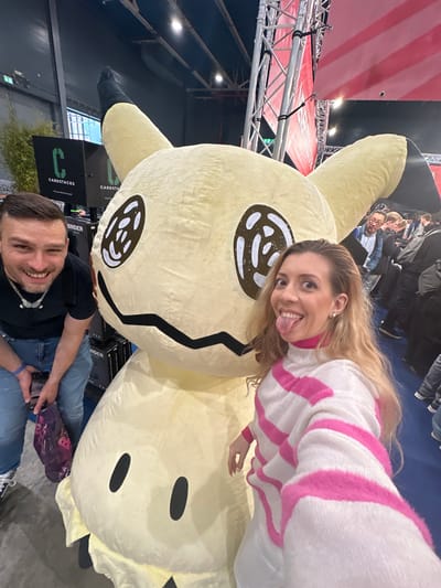 The image shows a group of people at an event, possibly a convention, in Utrecht, Netherlands. The main focus is on a large, plush Mimikyu Pokémon. A woman with long blonde hair and wearing a white and pink striped sweater is posing with the plush, sticking her tongue out. A man with short brown hair is seated near the plush, smiling and looking at his phone. Several other people are visible in the background. Signage for "CARDSTACKS" is visible near the lower left corner of the frame. A red and white striped banner is seen near the top left. The setting is indoors, with bright overhead lighting.
