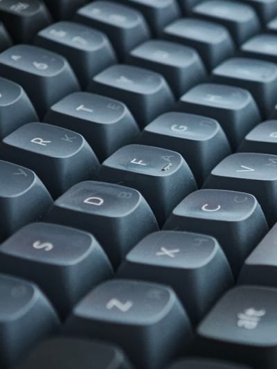 The image is a close-up, overhead shot of a black keyboard. The focus is sharp on the keys, revealing individual letters and symbols. The keys are arranged in a typical QWERTY layout, with rows of letters and numbers. Some of the visible keys include "S", "D", "R", "F", "X", "C", "V", "G", "A", "T", "2", and "alt". The lighting is dim, casting subtle shadows between the keys, suggesting an indoor setting, possibly at night or in a room with low ambient light. The shallow depth of field blurs the keys further back in the image, creating a sense of depth and drawing attention to the foregrounded keys. The overall aesthetic is clean and minimalist, with the dark color of the keyboard dominating the frame. There are no people or specific location cues other than the general context of a keyboard.