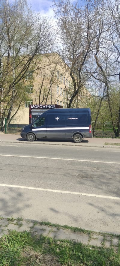 A blue van parked on the side of a road in Fryazino, Russia. The van has white stripes and lettering. The words "Мороженое" (ice cream) are printed vertically on the side of the van. The inscription "Спецсвязь" (Special Communications) is printed next to the emblem of the courier service of the Russian Federation. A building can be seen in the background through tree branches. There is a sidewalk and grass to the right of the road. The road is grey and has white lines painted on it.