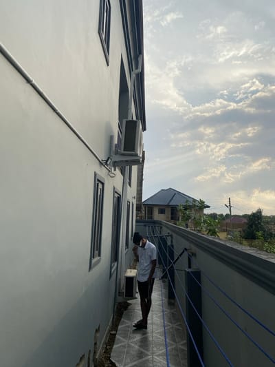 A low-angle perspective shows a light gray building with rectangular windows and an air conditioning unit mounted on the exterior. A man stands on a tiled balcony, looking at his phone. Overhead, a blue sky filled with wispy clouds is visible. The angle is disorienting, as the sky and building are tilted. In the background, a second building with a dark roof and green vegetation is partially visible.