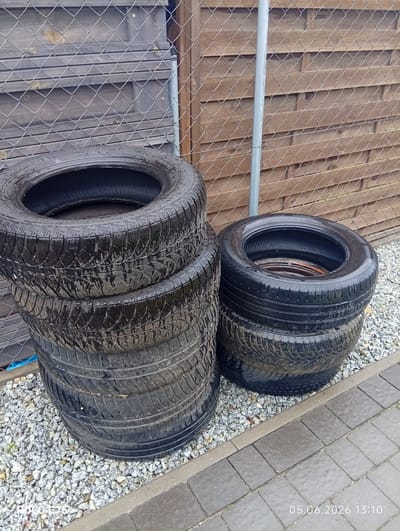 The image captures an outdoor scene featuring two stacks of wet car tires. A taller stack of five tires is on the left, with the topmost tire clearly displaying wet, dark rubber and tread patterns. To the right, a shorter stack consists of two similar wet tires. The tires rest on light-colored gravel, which covers the ground beneath and around them. Adjacent to the gravel on the right is an area paved with dark grey interlocking stones.

In the background, a chain-link fence spans the width of the frame, with a wooden slatted fence or wall visible behind it. A vertical silver metal pole, appearing to be a fence post, is situated between the two fence lines on the right side of the image. The wet surfaces of the tires suggest recent rain or damp weather conditions.

A digital timestamp in the bottom right corner of the image reads "05.06.2026 13:10," indicating the date and time the photo was taken. A watermark "POCO C75" is visible in the bottom left corner. The scene is located in Kiełczów, Poland. There are no people visible, and no specific activity is taking place beyond the static arrangement of the tires. Faint, unreadable text is present on the sidewalls of some tires.