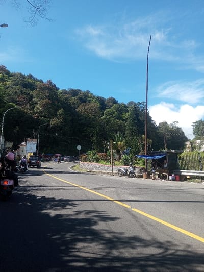 The image depicts a daytime scene on an asphalt road in a hilly, forested area, identified as Ka Thaung (Upper), Myanmar. The road, marked with a solid yellow center line, curves to the left in the mid-ground. Dense green and reddish-brown foliage covers a steep hillside dominating the left and background. The sky is bright blue with scattered white, wispy clouds.
In the immediate left foreground, the rear of a pink-hued scooter is visible, with a rider wearing a pink helmet. Additional scooters and a dark-colored car are observed further along the curving road. On the left, a white building with an ornate roof structure and a hanging banner is situated behind a streetlight pole.
On the right side of the road, a decorative stone wall borders a small planted area. A circular road sign is positioned within this area. Further right, a tall, slender pole extends vertically into the sky. Below this, a roadside stall operates under a blue tarp, with two individuals visible near it. One appears seated, the other standing. Various containers, including red and white buckets, are arranged around the stall. A white scooter is parked adjacent to the stall, and a green fence extends behind it. Shadows from overhead trees are cast across the road surface.