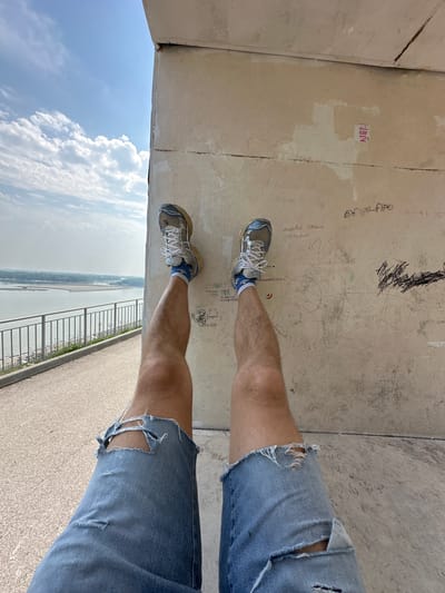 A first-person perspective shot shows a person's legs with ripped denim shorts and athletic shoes as they are suspended against a textured wall. The wall has graffiti and writing on it. To the left of the frame, a railing lines a path, with a body of water and a distant, hazy landscape visible beyond it. The sky above is blue with scattered clouds. The scene appears to be located at the Barnaul embankment in Barnaul, Russia, likely near the observation deck area.