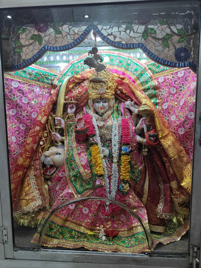 This image shows a statue of a Hindu deity, likely Durga, housed within a shrine in Gurgaon, India. The deity is depicted with multiple arms, adorned with elaborate golden jewelry, a crown, and richly colored, sequined clothing. The shrine itself is decorated with vibrant fabrics, including pink and green cloth with intricate patterns, and the background features a mosaic of reflective tiles and what appear to be floral motifs. To the left of the deity, a lion's head is visible, a common symbol associated with Durga. The statue is behind glass, suggesting it is part of a temple or a public display.