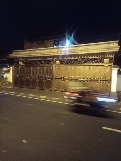 A night-time, eye-level, medium shot captures a wide, ornate, metal gate in Blahbatuh, Indonesia. The gate, comprised of multiple panels, is a muted gold color with intricate, dark scrollwork detailing and circular patterns. It is set against a dark background, with a building visible above. The gate appears to be part of a residential property. A white and yellow road marking runs along the asphalt road in front of the gate. A vehicle, possibly a motorcycle, is captured in motion, creating a blur of red and blue lights as it passes in front of the gate. The ambient lighting is low, suggesting it is late at night.