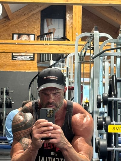 A man is taking a mirror selfie at a gym. The man has gray hair and a beard, is wearing a black baseball cap with "PORSCHE" in white letters, and a black tank top. The man's arms are heavily tattooed. In the background, there is weightlifting equipment, wood paneling, and framed pictures. The location could be in a gym located in the city of Jemelle, Belgium.