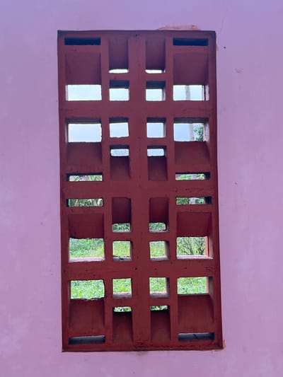 The image shows a terracotta decorative vent block embedded in a light pink wall. The vent block has a grid-like pattern of square and rectangular openings, through which a blurry view of green foliage and the sky can be seen. The wall appears to be weathered, with some cracks and imperfections visible. The setting is likely outdoors, given the visible vegetation. The style of the vent block suggests it could be found in older architecture or a more rustic environment. The image does not contain any people or specific identifiable landmarks that would pinpoint its exact location within Meteseh, Indonesia.