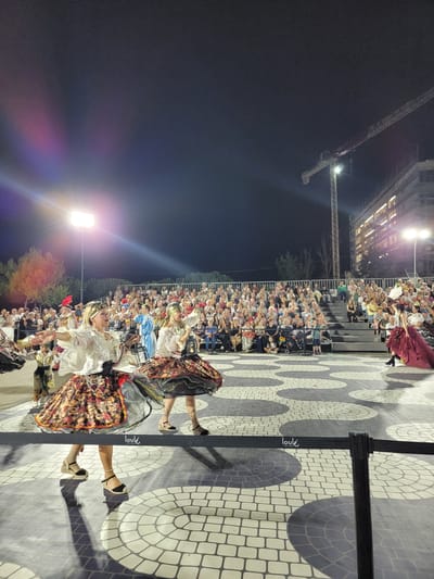 This image depicts an outdoor evening performance in Quarteira, Portugal, likely at the Largo do Mar or a similar public space. In the foreground, two women in traditional, colorful dresses are dancing, their skirts wide and voluminous, suggesting a lively folk dance. They are on a patterned paved area, with a black barrier in front of them. In the mid-ground, a large crowd is seated on tiered bleachers, watching the performance. Behind the audience, a construction crane and an illuminated building are visible against the night sky, indicating urban development in the area. The lighting suggests stage lights illuminating the performers and the audience, with a bright spotlight in the upper left corner. The overall scene captures a cultural event taking place in a public space in Quarteira, characterized by performance, audience engagement, and the backdrop of city infrastructure.