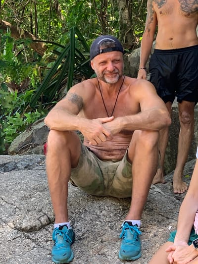 The image shows a man sitting on a rock in a jungle environment. He is wearing camouflage shorts and blue running shoes with white socks. He is shirtless and has a tattoo on his left arm. He is looking at the camera and smiling. Another person's legs and torso are visible to his right, wearing black shorts and standing. The background consists of lush green foliage and rocks. The scene appears to be outdoors in a natural setting, possibly a hiking trail or a waterfall area in Ko Samui, Thailand.