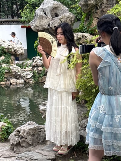 In a Chinese garden with a pond and rock formations, two young women are posing for a photograph. A woman on the right, wearing a blue dress with floral details and ruffles, is holding a camera and appears to be taking a photo of the woman on the left. The woman on the left, wearing a white dress and holding a fan, is looking down and to her side. In the background, another person wearing a hat and t-shirt is visible near a white building with a green door, suggesting the location is in Xicheng District, China. The garden features lush green foliage and natural rock landscaping.