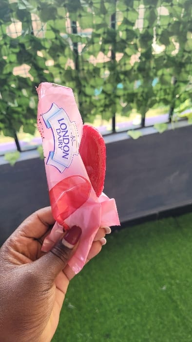 A dark-skinned hand, featuring a red and white tipped thumbnail, holds a partially unwrapped London Dairy brand red ice cream bar. The pink wrapper is torn open, exposing the crimson-colored frozen dessert. The background consists of a green artificial foliage screen, positioned above a dark horizontal railing or surface. Below this, an area of artificial green turf is visible. The scene is illuminated by natural daylight. No specific landmarks or unique environmental features are present within the image to definitively identify a precise location in Nanyuki, Kenya.