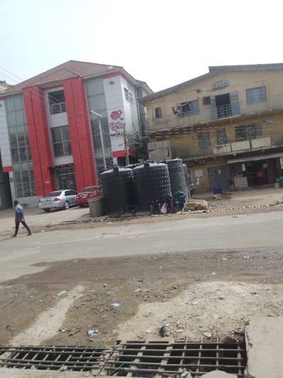 The scene depicts a daytime street view in Sabon Gari West, Nigeria, characterized by urban infrastructure and mixed-use buildings. In the foreground, a paved road shows scattered patches of moisture and dirt, leading to a shoulder of uneven dirt and debris. A metal drain grate occupies the bottom right.

On the left, a modern three-story building with a red tile roof features a white and red facade, incorporating large glass panels and prominent red vertical cladding. A sign on its front displays "PR Royal Resort." Two cars are parked on the adjacent road: a silver sedan and a red sedan.

To the right of the modern building, an older, three-story building with a light beige, weathered facade stands. Its upper floors have multiple windows, some fitted with metal security bars or grates, and visible balconies with clothes hanging. The ground floor houses several small, open-fronted shops or stalls where a few individuals are partially visible.

Centrally located between the road and the older building, a cluster of approximately six large, black plastic water storage tanks is positioned on the dirt shoulder. Several individuals are seated or gathered around these tanks, some partially obscured. One person is distinctly visible wearing a blue garment.

A single pedestrian, wearing a light-colored top and dark trousers and carrying a bag, walks from left to right on the paved road in the left-midground. Overhead, a utility pole with wires is positioned in front of the modern building, near the water tanks. The sky is bright and clear, indicating daylight conditions.