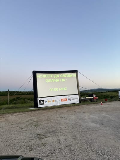 The image shows an inflatable outdoor movie screen set up on a gravel surface. The screen displays the text "МОЖЕТЕ ДА СЛУШАТЕ ФИЛМА НА : 99.00 MHZ", which translates to "You can listen to the movie on: 99.00 MHZ". Several sponsor logos are visible along the right side of the screen: "Evolution", "STIHL", "MOTO-PFOHE rent a car", and "Petrol". A power line is visible in the middleground and trees are shown in the background. A red cone is visible in the upper right of the image, and a truck is behind the screen. The sky indicates it is dusk. The location is Lukovit, Bulgaria.
