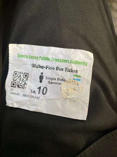 A close-up image shows a worn, crumpled white paper bus ticket lying on a dark, possibly fabric surface. The ticket, issued by the "Sierra Leone Public Transport Authority," is identified as a "Waka-Fine Bus Ticket" for a "Single Ride" and a "General" fare, indicated by a silhouette of a person. The cost displayed is "Le. 10." A circular gold holographic sticker, featuring "SLPTA" and "WAKA-FINE BUS TICKETS" repeated around its edge, is affixed to the ticket's upper right. A QR code is present on the lower left, accompanied by the alphanumeric codes "65 GMA40" and "M8T6-GMA40." The crumpled condition suggests the ticket has been used. This item points to public transportation in Freetown, Sierra Leone, likely a local bus service.