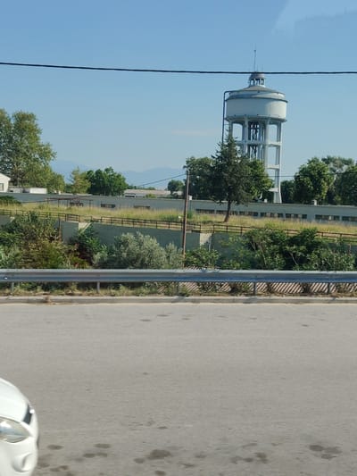 This image depicts a water tower in Larissa, Greece, on a clear day with a blue sky. The water tower is a tall, cylindrical structure with a domed roof, supported by a multi-legged concrete base. Surrounding the water tower are various trees and vegetation, and in the foreground, a road and a guardrail are visible. The scene appears to be a street-level view, possibly from a vehicle, as suggested by the angle and the presence of a car headlight in the lower-left corner of the frame. The overall atmosphere is serene and rural, with the water tower standing as a prominent landmark against the backdrop of the natural landscape and the distant mountains.