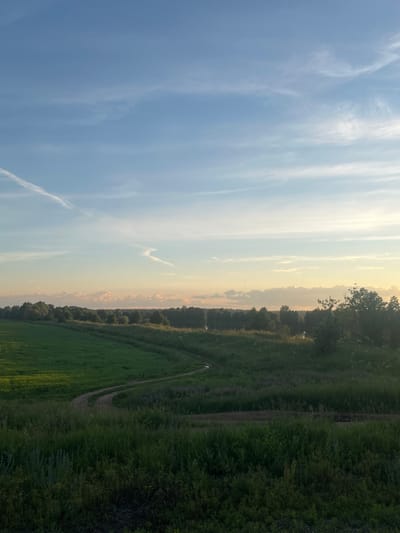 The image depicts a serene landscape with a winding dirt path cutting through lush green fields. Tall trees line the horizon, interspersed with what appears to be a small body of water in the distance. The sky above is a gradient of soft blues and warm oranges, with wispy clouds streaking across the expanse. The overall impression is one of natural beauty and tranquility, characteristic of a rural setting. While the image does not provide explicit details about Kauchuk Stantsiya, Russia, it evokes a sense of peaceful countryside often found in such locations.