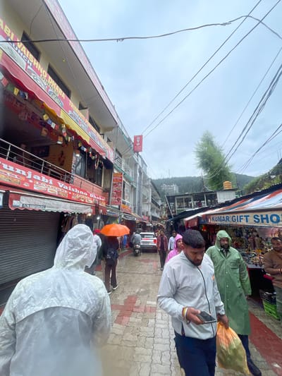 This is a street scene in Bhagsu, India, on a cloudy day. The street is paved with reddish-brown and grey tiles. There are several buildings lining the street, with shops on the ground floor and more levels above. One building prominently features signs for "Punjabi Vaishno Dhaba" and "Trimurti Punjabi Vaishno Dhaba," indicating it is a restaurant. Another shop is labeled "Kumar Gift Shop."

Several people are walking along the street, many wearing raincoats. One person in the foreground is wearing a white raincoat and has a patterned design on the back. Another person in a green raincoat is walking with a grocery bag. An orange umbrella is visible as someone walks away from the camera. A white car is parked on the street, and a motorcycle is also present. The background shows a hillside covered with trees and some buildings. Overhead, power lines crisscross the sky.