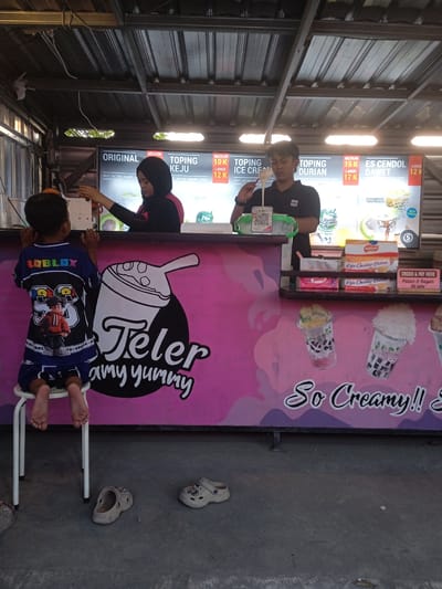 In Punden, Indonesia, a child is seated on a white stool in front of a food stall. The child, wearing a blue and black Roblox-themed outfit, faces away from the camera, observing the stall. Behind the child, a woman in a black hijab and a pink shirt is attending the stall, interacting with another person who is not fully visible. The stall features a pink counter with various items displayed, including signage for "Teler & Ice Cream" and product promotions. The background reveals a metal roof structure and a tiled wall. On the ground, in front of the stall, two pairs of sandals are visible. The overall scene depicts a bustling street food vendor environment.
